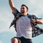 A joyful young man jumps midair with clouds and blue sky in the background, exuding energy and freedom.
